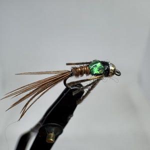 May include: A fishing fly with a green bead head, brown body, and brown feather tail. The fly is held by a black tool, possibly a fly tying vise. The fly is designed to imitate an insect, used for fly fishing.