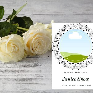 Puede incluir: Una tarjeta conmemorativa con un marco circular decorativo, una ilustración de paisaje y el texto "In Loving Memory of Janice Snow, 15 August 1945 - 10 May 2025." Dos rosas blancas y hojas verdes sobre una superficie de madera blanca.