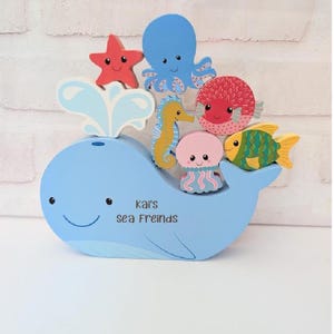 May include: A light blue wooden whale with a smiling face and the text "Kai's sea Freinds". The whale has a water spout and is surrounded by colourful wooden sea creatures, including a starfish, octopus, seahorse, and fish.