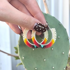 May include: A pair of hoop earrings featuring a gradient of small beads in cream, yellow, orange, and red, with a small turquoise and black geometric accent at the bottom. The beads are arranged in a circular pattern on a thin silver-toned metal frame.