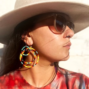 May include: A person wearing a wide-brimmed hat and sunglasses with large, colorful beaded hoop earrings. The earrings are made of multiple layers of beads in shades of red, orange, yellow, blue, and black.