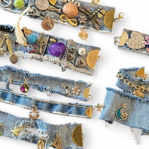 May include: Several denim bracelets with various charms and embellishments. The charms include coins, keys, hearts, and other decorative elements. The bracelets have gold-colored clasps and are arranged on a white surface.