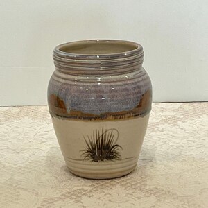 May include: A handcrafted ceramic jar with a wide opening and a slightly textured surface. The jar features a cream-colored base with a painted design of a cluster of grass. The upper portion has a band of purple, brown, and tan hues.