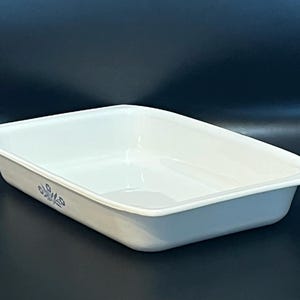 May include: A rectangular white ceramic baking dish with rounded corners and a smooth interior. The exterior side features a small, delicate blue floral pattern consisting of three stylized flowers on thin stems with green leaves.