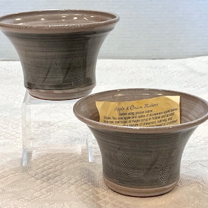 以下が含まれることがあります： 手作りのリンゴと玉ねぎ用のセラミックベーカリー2つ。グレーのベーカリーは波模様と薄茶色の縁があります。1つのベーカリーには、テキストが書かれた小さな紙タグが内側にあります。ベーカリーは透明なアクリルスタンドにあります。