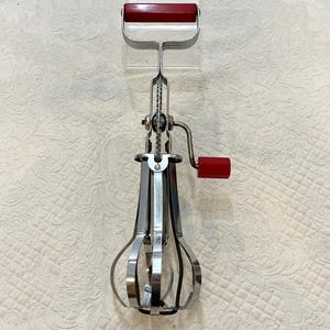May include: A vintage hand-crank egg whisk with a red handle and crank. The whisk is made of shiny metal and features a gear mechanism. The handle is rectangular and the crank handle is cylindrical. The whisk's cage is a series of curved metal loops.