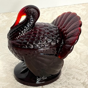 May include: A ruby-red glass turkey figurine with a textured body and fanned tail. The turkey has a curved neck and a small, round base. This decorative piece is likely a candy dish or trinket box.