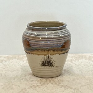 May include: A ceramic vase with a wide, round body and a slightly flared rim. The vase features horizontal bands of color, including brown, blue, and cream. A stylized plant design is painted on the lower portion of the vase.
