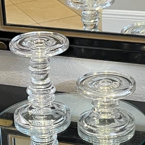May include: Two clear glass candle holders with a tiered, circular base and a matching circular top platform. The taller holder features a multi-ringed, stacked stem design, while the shorter holder has a more compact, single-ringed stem.