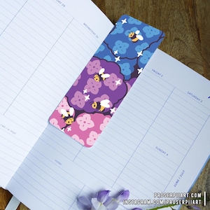 May include: A purple, pink, and blue bookmark with a repeating pattern of bees and flowers. The bookmark is made of sturdy cardstock and is perfect for marking your place in a book.