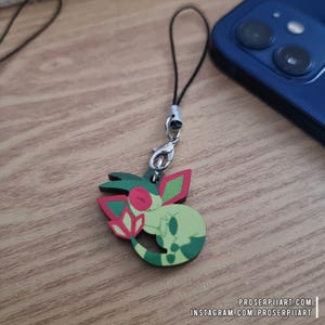 Puede incluir: Un colgante de madera con un personaje de dibujos animados verde y rojo, sujeto a un cordón negro con un cierre plateado. El colgante está sobre una superficie de madera cerca de un smartphone azul. El colgante tiene el texto "proserpiiart.com" e "instagram.com/proserpiiart".