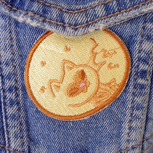 Op de afbeelding: Een geborduurde patch in geel en oranje van een kat die slaapt op een halve maan. De patch is genaaid op een blauw denim jack.