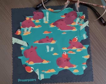 Puede incluir: Un paño de limpieza cuadrado de microfibra con un fondo turquesa que presenta hipopótamos de dibujos animados en un estanque con pájaros. El paño tiene un borde azul oscuro y el texto "ProserpiiArt". Gafas con montura dorada descansan sobre el paño.