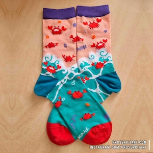Può includere: Un paio di calzini colorati con un design a tema granchio e oceano. I calzini presentano granchi rossi su uno sfondo color pesca con onde turchesi e un polsino viola. La punta e il tallone sono rossi e il testo "prose" è visibile.