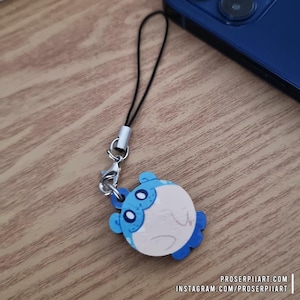 May include: A wooden charm featuring a cartoon pufferfish design in white, light blue, and dark blue. The charm is attached to a black lanyard with a silver-colored clasp. The charm is lying on a wooden surface. The text "proserpiiart.com" and "instagram.com/proserpiiart" are visible.
