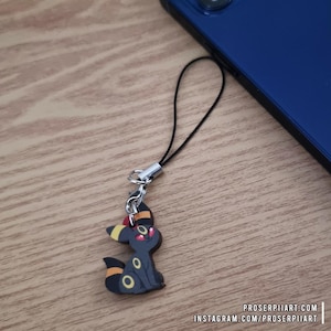 Può includere: Un ciondolo Umbreon nero e giallo con un cordino nero e una chiusura color argento. Il ciondolo è una figura in stile cartone animato con dettagli gialli e occhi rossi. Il ciondolo è accanto a un telefono blu su una superficie di legno. Il testo "PROSERPIIART.COM" e "INSTAGRAM.COM/PROSERPIIART" è visibile.