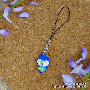 Può includere: Un ciondolo a forma di pinguino blu e bianco con un viso sorridente e ali spiegate, attaccato a uno stringa nera con un fermaglio argentato. Il ciondolo è su uno sfondo di sughero marrone con fiori viola.
