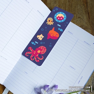 May include: A bookmark with a dark blue background featuring a colorful octopus, a fish, and a jellyfish. The bookmark also has a circle with a cat face inside.