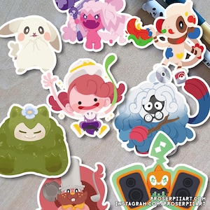 Op de afbeelding: Een verzameling kleurrijke cartoonkarakterstickers. De stickers hebben verschillende ontwerpen, waaronder een wit konijn, een roze wezen, een groen wezen met een bloem en een blauwharig personage. De stickers liggen op een grijze ondergrond.