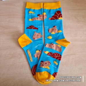 Op de afbeelding: Een paar blauwe sokken met een gele boord en een patroon van capibara's die zwemmen in een zwembad met gele rubberen eendjes. De sokken hebben de tekst "PROSERPI" op één sok.