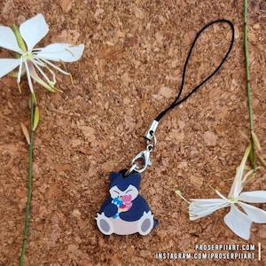 Könnte beinhalten: Ein marineblauer und beigefarbener Anhänger mit einem Cartoon-Snorlax-Charakter, der bunte Leckereien hält. Der Anhänger ist an einer schwarzen Kordel mit einem silberfarbenen Verschluss befestigt. Weiße Blumen und Text befinden sich im Hintergrund.