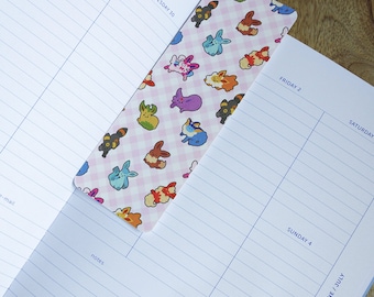Op de afbeelding: Een roze en witte gingham-bladwijzer met een herhalend patroon van cartoon Pokémon-personages. De bladwijzer is geplaatst op een pagina van een planner met de data "Woensdag 30", "Vrijdag 2", "Zaterdag" en "Zondag 4" die op de pagina staan.