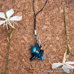 Può includere: Un ciondolo nero e blu raffigurante un personaggio dei cartoni animati con dettagli gialli e blu. Il ciondolo è attaccato a un cordino nero con un fermaglio color argento. Lo sfondo è una superficie marrone strutturata con fiori bianchi e steli verdi.