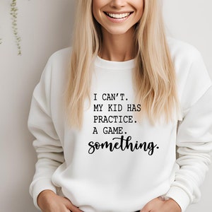 May include: White sweatshirt with the text "I can't. My kid has practice. A game. something." in black script. The sweatshirt is a crew neck style and appears to be made of a soft material.