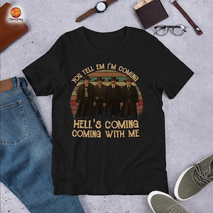 May include: Black t-shirt featuring a vintage-style graphic of four men in Western attire against a sunset backdrop. The text reads "YOU TELL EM I'M COMING" and "HELL'S COMING COMING WITH ME".