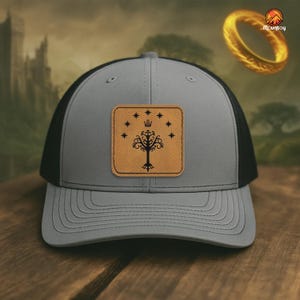 May include: Grey and black trucker cap with a leather patch displaying a tree and stars design. The cap has a curved peak and a mesh back. The patch is brown with black detailing. A classic accessory.