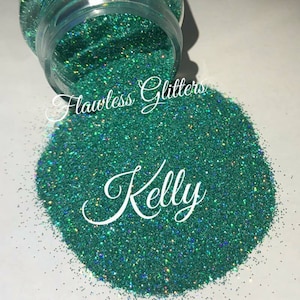 Op de afbeelding: Een close-up van een pot met teal groene glitter met een iriserende glans. De glitter stroomt uit de pot en de tekst "Flawless Glitters Kelly" is zichtbaar op de afbeelding.