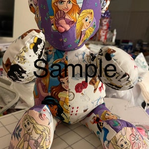 May include: A purple and white patchwork teddy bear with a variety of Disney princess characters on the fabric. The bear is sitting on a white and purple checkered surface. The word "Sample" is written on the bear's chest.