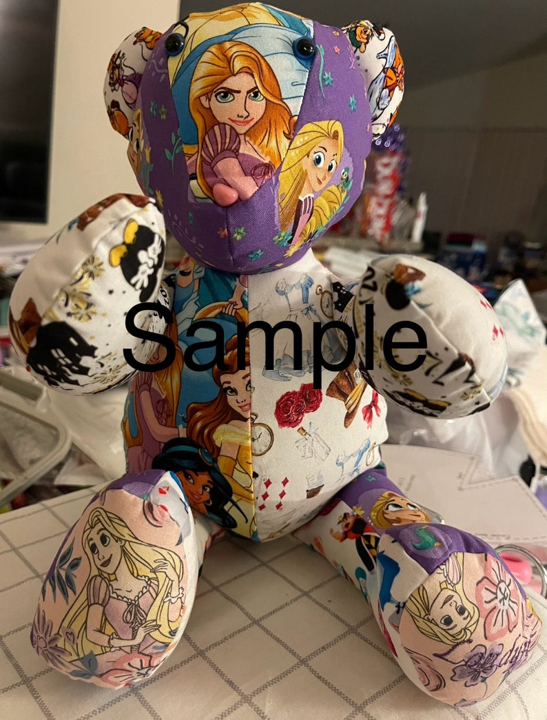 May include: A purple and white patchwork teddy bear with a variety of Disney princess characters on the fabric. The bear is sitting on a white and purple checkered surface. The word "Sample" is written on the bear's chest.