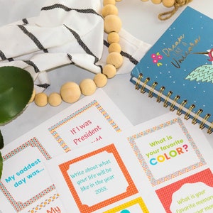 May include: A set of colorful cards with prompts for a game or activity. The cards have a variety of questions, such as "What is your favorite color?" and "My saddest day was..." The cards are on a white surface with a wooden bead garland and a blue notebook with a unicorn design.