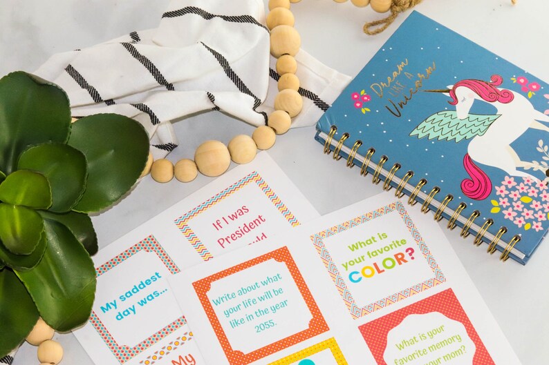 May include: A set of colorful cards with prompts for a game or activity. The cards have a variety of questions, such as "What is your favorite color?" and "My saddest day was..." The cards are on a white surface with a wooden bead garland and a blue notebook with a unicorn design.