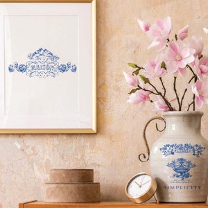 Può includere: Un'anfora in ceramica bianca con disegni floreali blu e le parole "Blessed" e "Simplicity" sul davanti. L'anfora è piena di fiori di magnolia rosa e si trova su un tavolo di legno con un orologio dorato e due scatole rotonde marroni.