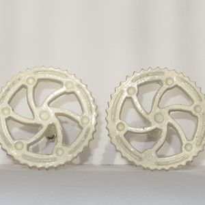 May include: A pair of white metal decorative drawer pulls shaped like a bicycle gear with a swirl design.