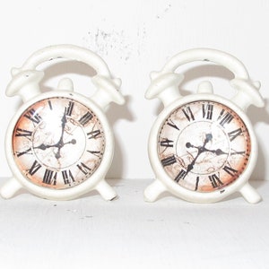 May include: Two white ceramic alarm clocks with black Roman numeral faces and black hands. The clocks have a distressed, vintage look.