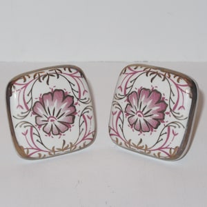 Puede incluir: Dos tiradores de cajón cuadrados de cerámica con un borde plateado. Los tiradores presentan un diseño floral rosa con detalles dorados sobre un fondo blanco.