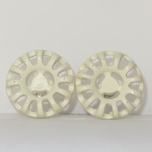 May include: Two off-white cast iron decorative knobs with a circular design and a central hole. The knobs have a raised, sunburst-like pattern around the edge and a smaller, raised, teardrop-shaped design in the center.