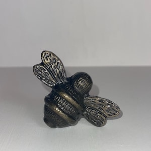 May include: A bronze bee-shaped cabinet knob with a textured finish. The bee has a segmented body with a gold stripe around the middle.