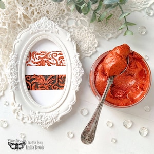 Może przedstawiać: Zbliżenie na mały słoik z metaliczną pomarańczową farbą do prac ręcznych, z łyżką nabierającą trochę farby. Farba jest gęsta i ma metaliczny połysk. Słoik stoi na białym tle z ozdobną białą koronką i przezroczystymi szklanymi kamieniami.