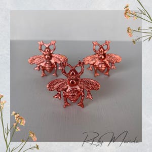 May include: Three rose gold-coloured bee-shaped decorative objects. The bees have detailed wings and body segments. The objects are arranged on a light grey surface, with floral accents in the corners.