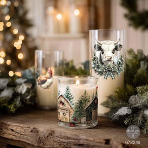 Op de afbeelding: Drie glazen kandelaars met feestelijke ontwerpen. Eén toont een haan, een andere een koe met een krans en een geruite strik, en de voorste toont een besneeuwd schuurtje met dennenbomen en cadeautjes. Elke houder bevat een witte kaars.