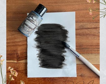 May include: A bottle of black liquid acrylic ink next to a white paper with a black ink swatch and a paintbrush. The ink bottle is labeled "Ink Black" and "Liquid Acrylic". The background is a wooden surface.