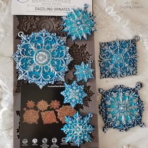 Könnte beinhalten: Eine Silikonform zum Herstellen von sechs verschiedenen Schneeflocken-Ornamenten. Die Form ist 12,7 cm x 25,4 cm groß und besteht aus 100 % Silikon. Die Form ist backofengeeignet, mikrowellengeeignet und gefriergeeignet. Die Form ist außerdem lebensmittelecht und antihaftbeschichtet. Die Form wird mit fertigen Ornamenten in Blau und Silber gezeigt.