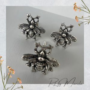 Może przedstawiać: Trzy srebrne gałki do szafek w kształcie pszczół. Gałki mają szczegółowe skrzydła, segmenty ciała i czułki. Gałki znajdują się na jasnoszarej powierzchni. Gałki są prawdopodobnie przeznaczone do mebli lub dekoracji.