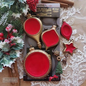 May include: A gray silicone mold set labeled Noel Elegance featuring various Christmas ornament shapes. The set includes a large circular mold, two teardrop-shaped molds, and a star-shaped mold, all filled with a vibrant red material.