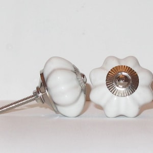 Peut inclure: Deux boutons de cabinet en céramique blanche avec un design cannelé. Un bouton a un accent en métal argenté au centre.