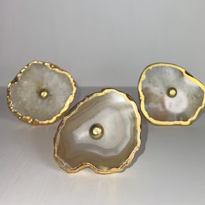 Puede incluir: Tres tiradores de cajón de ágata con ribete dorado. Cada tirador presenta un patrón único de ágata blanca y marrón.